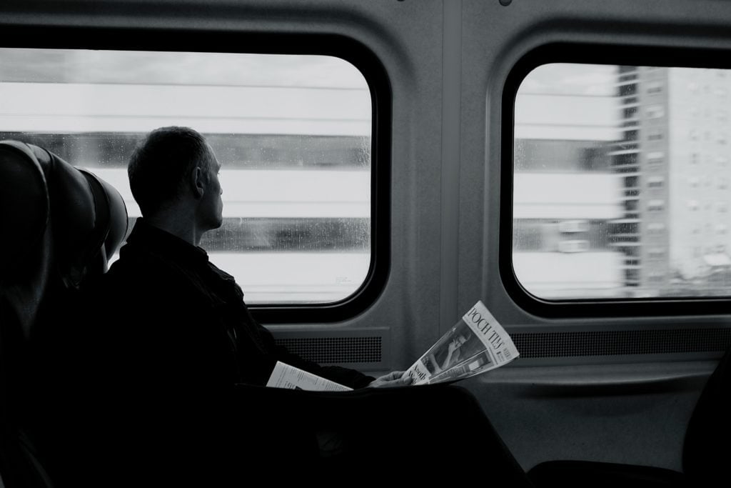 ein Mann der in einem Zug sitzt und aus dem Fenster schaut während er eine Zeitung in seinen Händen hält.