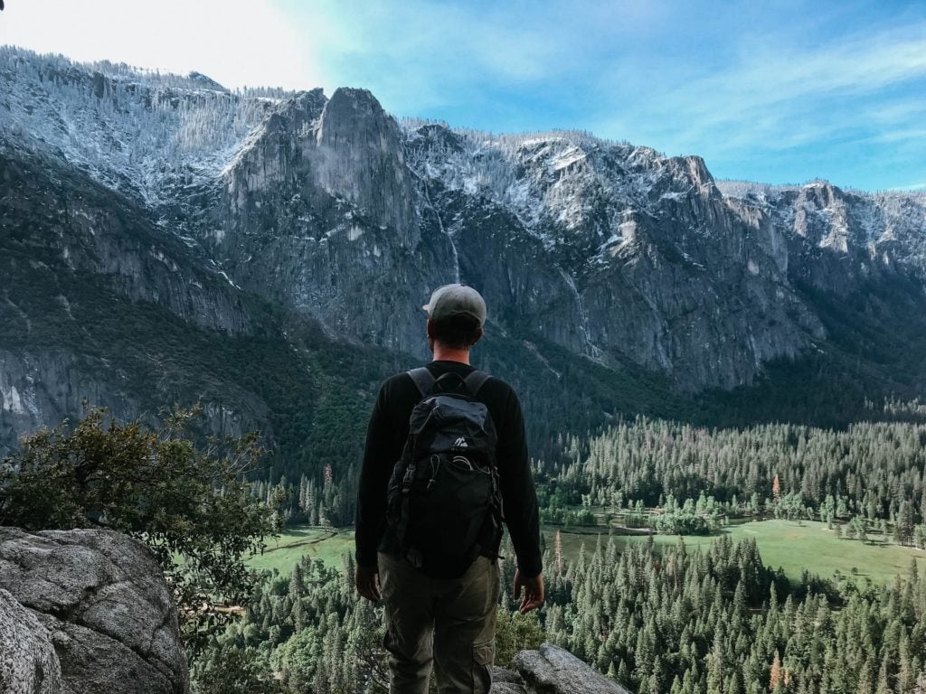 Mann mit Mütze und Rucksack von hintem auf grünem Untergrund in den Bergen, im Hintergrund wolkiger Himmel 