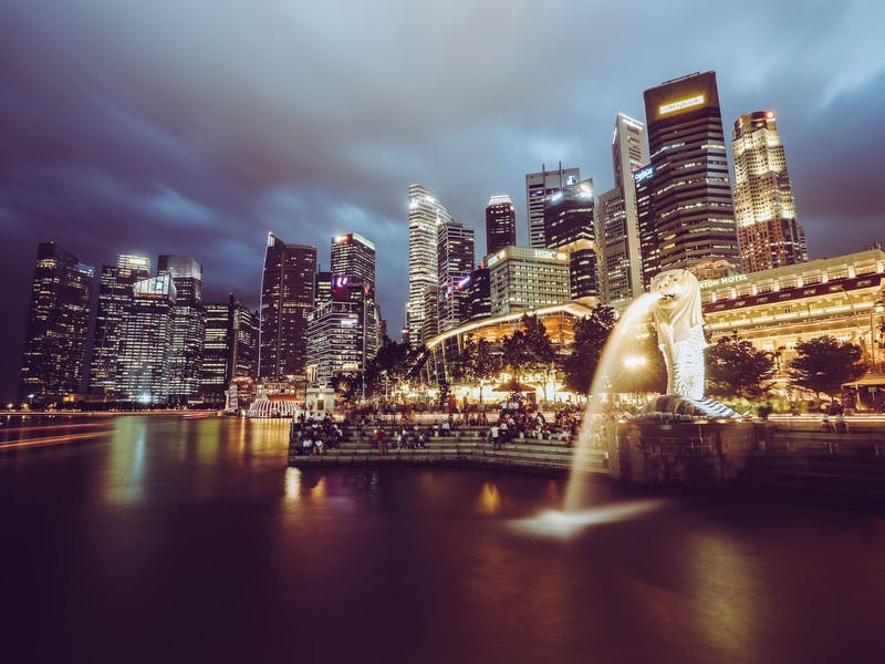 Der Hafen von Singapur bei Nacht, mit dem Löwen-Wahrzeichen im Vordergrund und der hohen Skyline im Hintergrund 