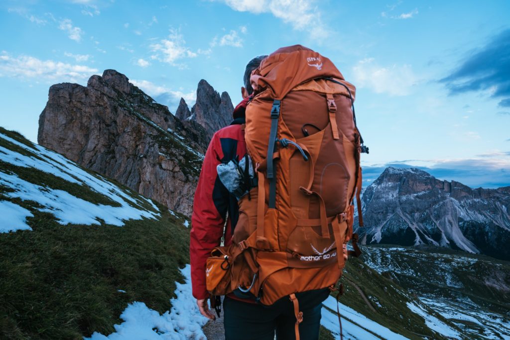 Mann in roter Jacke mit orangenem Backpack steht auf teils schneebedecktem Abhang, im Hintergrund Berge