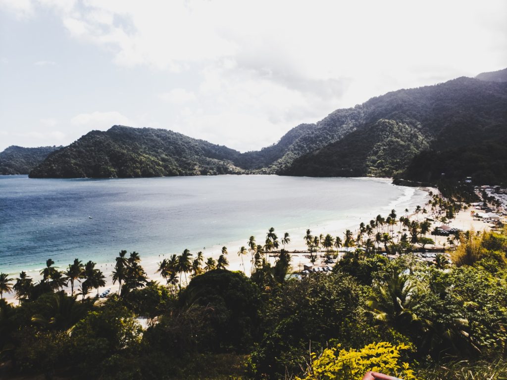 a view of the calm body of water by a white sandy beach and a view of the green landscapes with mountains and trees in the best caribbean islands of Trinidad & Tobago