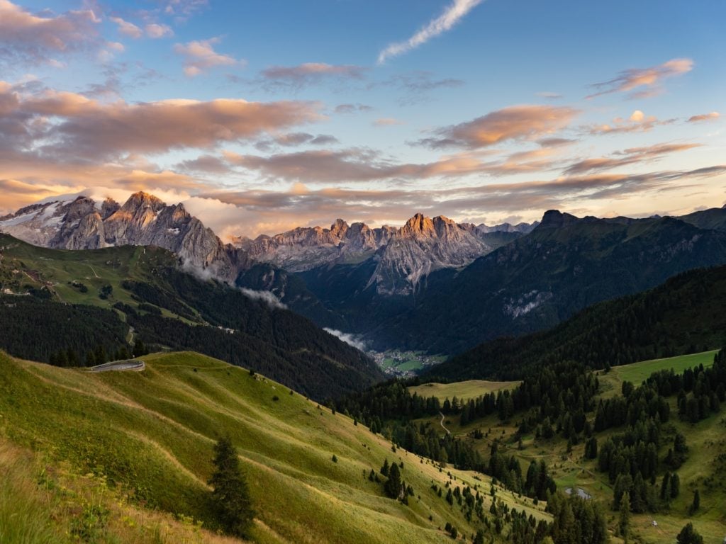 green grass fields and trees, surrounded by big mountains under white clouds and blue skies in Sella-Herbetet Traverse, Italy, one of the best alpine hiking trails
