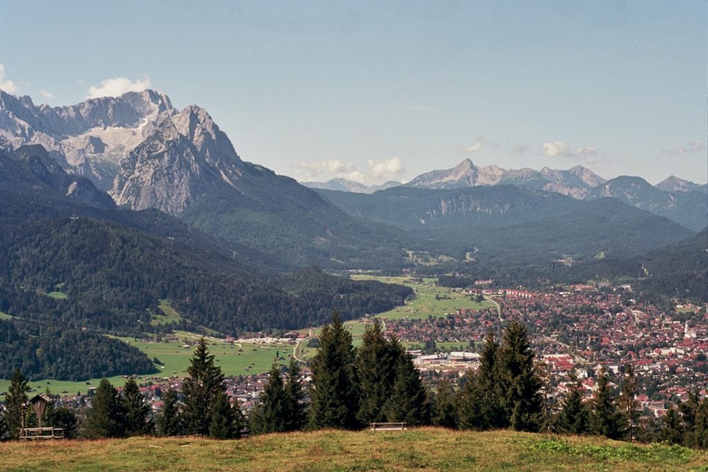 daytime photo of the landscape of Garmisch-Partenkirchen showing the mountains and the town below it surrounded by nature and greenery, the perfect hiking trail