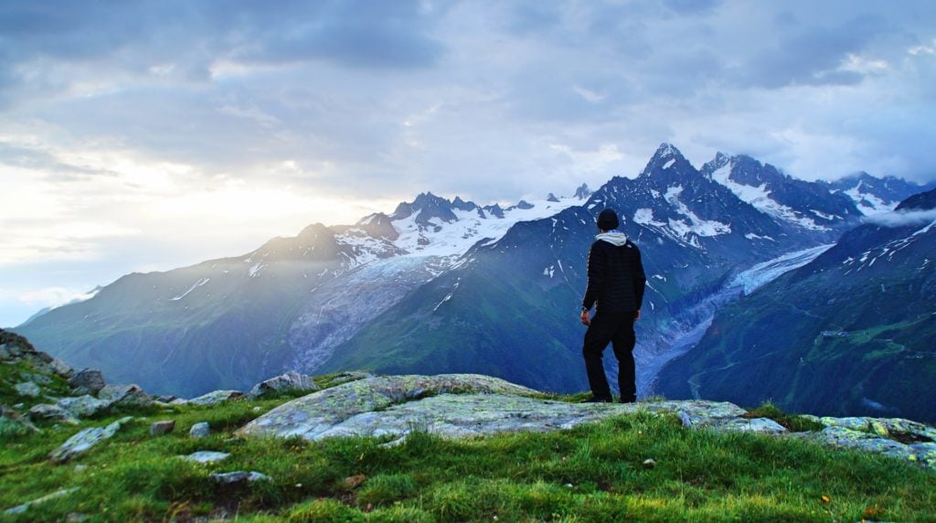 person standing on green grass while looking over a beautiful landscape in the french alps, one of the best alpine hiking trails in Chamonix France